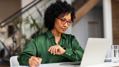 A woman in glasses writing notes while working on a laptop at a desk.