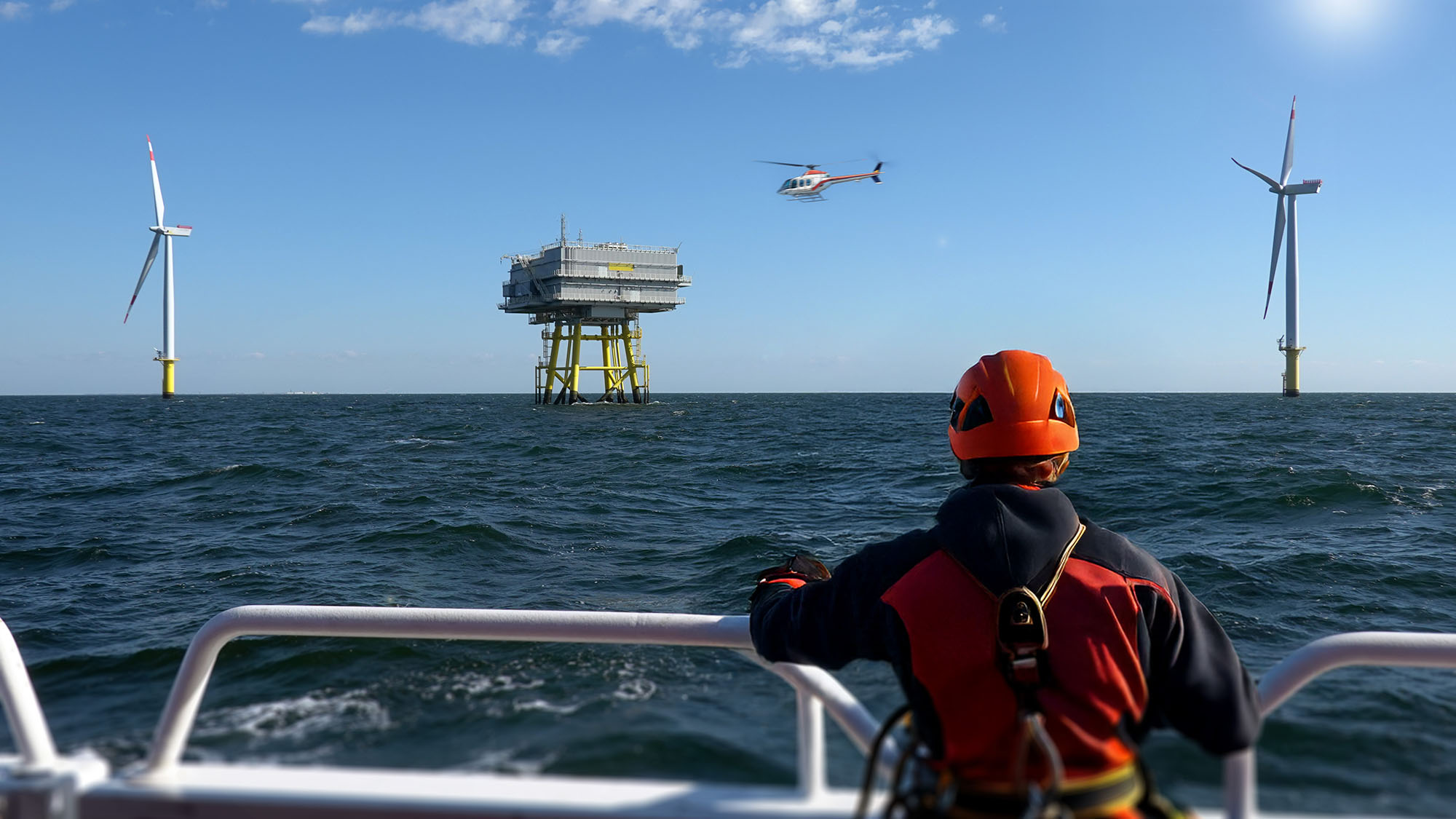 Man dressed in red safety gear standing on a boat looking at a wind farm as a helicopter lands on an offshore platform 
